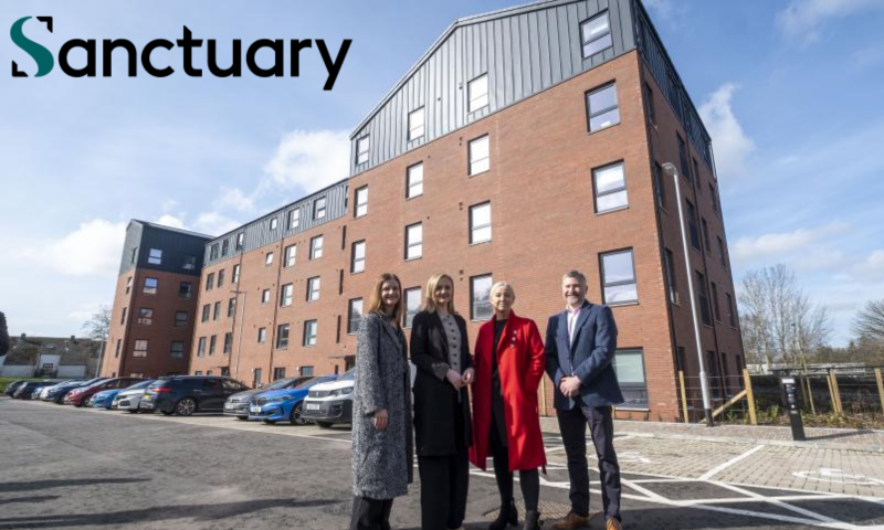 This image shows a wide-angle, outdoor shot of four people standing in front of a modern, multi-story apartment building.  The Setting The building is a large, red-brick structure with five floors. It has a distinctive architectural style:  The top section and some side panels are clad in dark grey or black corrugated metal.  The windows are uniform, framed in dark grey, and reflect the bright daylight.  The building sits behind a paved parking area with several cars (blue, white, and dark grey) parked in a row to the left.  The ground in the foreground is asphalt, marked with white lines for parking spaces, including a designated disabled parking bay.  The People Four people are standing together in the center-right of the frame, positioned as if posing for a professional or promotional photograph. From left to right:  A woman wearing a long, grey patterned wool coat and black trousers.  A woman in a long black coat worn over a beige top and black trousers.  A woman wearing a vibrant, knee-length red coat over dark clothing, which stands out as the brightest element in the image.  A man in a navy blue blazer, a light-colored collared shirt, and dark trousers.