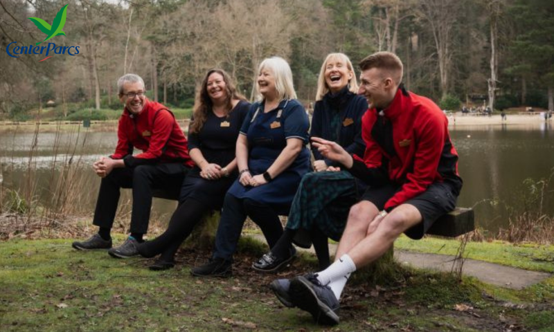 Gemini said Five people are sitting on a wooden bench outdoors, with a lake and a dense forest in the background. The group consists of three women and two men, all appearing to be in a lighthearted and joyful mood, with several of them laughing or smiling broadly.  The two men are wearing bright red and black zip-up jackets and black shorts or trousers, while the women are dressed in navy blue and dark-colored clothing. Some members of the group are wearing name tags, suggesting they may be colleagues. They are positioned on a grassy area near the water's edge, and the overall atmosphere is one of camaraderie and relaxation in a natural setting.