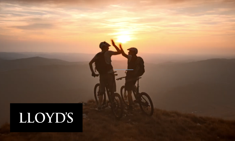 The image shows two mountain bikers in silhouette, high-fiving on a hill at sunset. They are standing with their bikes, looking out over a valley. The sun is setting behind them, creating a warm, golden glow. The Lloyd's logo is in the bottom left corner.