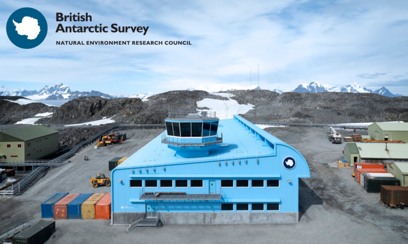 The image shows the Discovery Building at the Rothera Research Station in Antarctica. It is a large, two-storey modern facility characterised by its striking bright blue exterior, designed to stand out against the stark, monochromatic polar landscape.  Architectural Details Structure: The building has a long, aerodynamic shape with a distinct curved roofline on one side, which serves as a wind deflector to prevent snow build-up.  Control Tower: A multi-sided, glass-walled control tower sits atop the flat section of the roof, providing a 360-degree view of the station and the surrounding runway and wharf areas.  Windows: Rows of rectangular windows line the upper and lower floors, designed to maximise natural light while maintaining high thermal efficiency.  Foundation: The building is set on a solid concrete plinth, elevating the metal-clad structure slightly above the rocky, permafrost ground.  Surroundings and Context Landscape: The building is situated in a rugged, rocky environment with sparse patches of snow. In the background, jagged, snow-capped mountains and glaciers rise under a pale blue, slightly cloudy sky.  Logistics: To the left and right of the building, several colourful shipping containers (blue, orange, and red) and heavy machinery, including a yellow telehandler, indicate ongoing operational and construction activity.  Branding: The British Antarctic Survey (BAS) logo—a circular emblem featuring a map of Antarctica—is prominently displayed on the side of the building.  The overall impression is one of cutting-edge engineering and resilience, a high-tech "hub" sitting in one of the most remote and extreme environments on Earth.