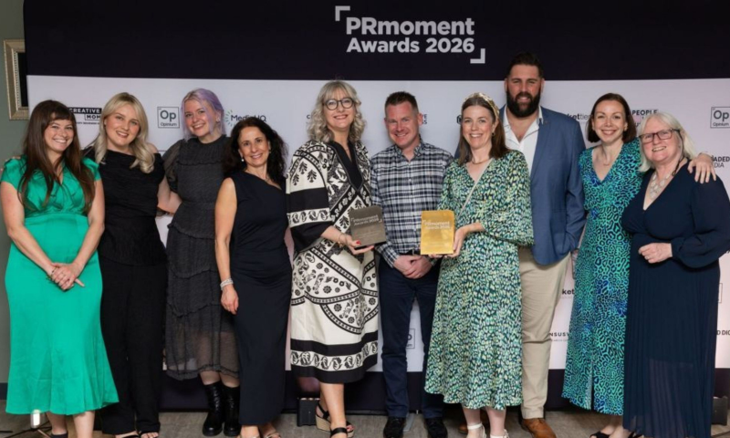 The Group: The group consists of seven women and three men, all dressed in smart-casual or formal attire. Their expressions are joyful and smiling, suggesting a celebratory atmosphere.  The Awards: Two individuals in the center of the group are prominently holding awards. One is a dark, rectangular plaque held by a woman in a patterned black-and-white dress, and the other is a gold-colored rectangular award held by a woman in a green patterned dress.  Setting: The photo is taken against a professional "step and repeat" banner. Bright studio lighting is visible on the floor at the edges of the frame, and the floor appears to be a light-colored wood or laminate.  Fashion: The attire varies from vibrant colors, such as a bright green dress and a blue leopard-print dress, to classic black outfits and a blue blazer.