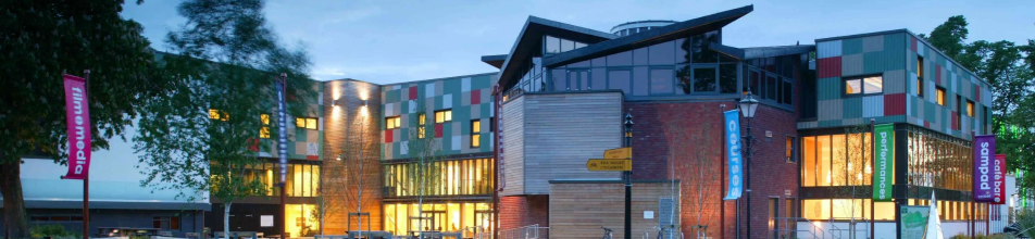 Architectural Features The Facade: The building is characterized by a distinctive multi-coloured "pixelated" cladding on the upper levels, featuring a palette of green, grey, cream, and red rectangular panels.  Glazing: The ground floor features extensive floor-to-ceiling glass curtain walls, allowing warm light from the interior—likely the foyer and café areas—to spill out onto the courtyard.  Central Structure: A prominent central section features horizontal timber slats and a striking, angular sawtooth roofline, which adds a dynamic geometric profile to the complex.  Materials: The design utilises a mix of traditional red brick, natural wood, and modern aluminium cladding, reflecting the centre's evolution since its original opening in 1964.  Signage and Banners Several tall, vibrant vertical banners line the walkway, highlighting the Midlands Arts Centre's diverse cultural offerings:  Film & Media: A pink banner on the far left.  Courses: A bright blue banner near the centre.  Performances: A green banner towards the right.  Sampad: A purple banner referencing the South Asian arts organisation based at the MAC.  Café Bar: A red banner on the far right.
