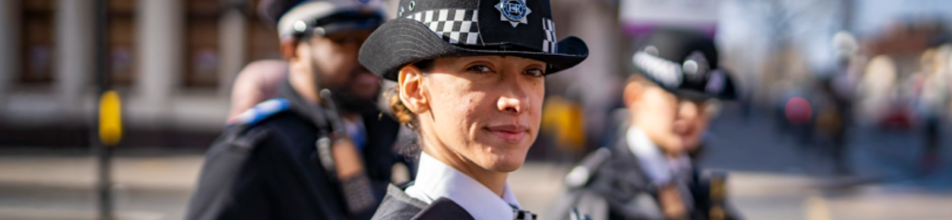 This is a wide, landscape-oriented photograph focussing on a female police officer, with two other officers visible in the blurred background.  Here are the key details of the image:  Foreground Subject: A woman looking directly toward the camera with a subtle, confident smile. She is wearing a traditional British police uniform, including a white collared shirt, a dark jacket, and a distinctive bowler-style police hat featuring the black-and-white chequered Sillitoe tartan band and a police badge on the front.  Background Subjects: To her left (the viewer's left), a male officer is visible in profile, looking off to the side, wearing a peaked police cap and a uniform equipped with a radio. To her right, another officer is partially visible and also blurred, wearing a similar bowler-style hat.  Setting: The scene is outdoors on what appears to be a city street during the daytime. The background elements—including buildings, streets, and distant figures—are softly out of focus, creating a shallow depth of field that emphasises the central officer. Bright, natural sunlight illuminates the side of her face.