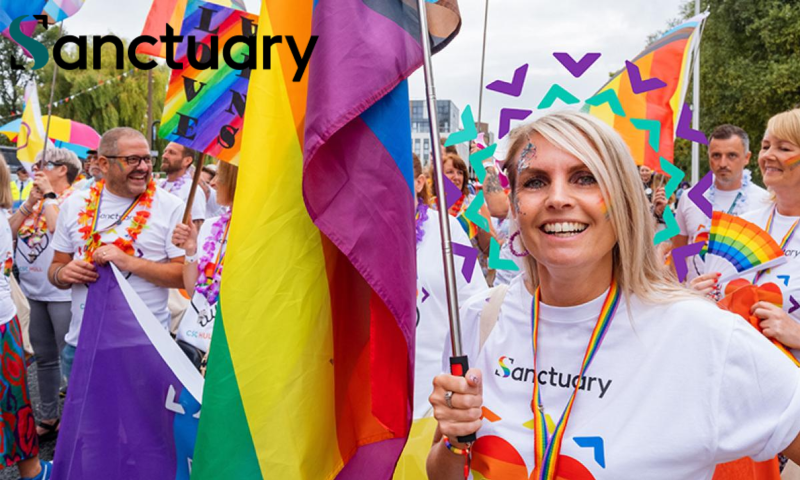 The image captures a vibrant, cheerful group of people participating in an outdoor Pride parade or festival. The atmosphere is celebratory, filled with rainbow colors, smiles, and a sense of community.  Key Elements Foreground Subject: The central focus is a blonde woman smiling warmly at the camera. She has festive rainbow face paint on her cheek and glitter near her eye. She is holding a large rainbow flag on a pole.  Attire: Most participants, including the main woman, are wearing matching white T-shirts that feature a rainbow heart logo and the word "Sanctuary" written across the front. The woman also wears a matching rainbow lanyard.  The Crowd: Surrounding her are several other smiling participants of varying ages. To her left, a man with a rainbow flower lei and glasses laughs heartily. To her right, a woman holds a folded rainbow hand fan.  Flags and Signs: Multiple flags are visible throughout the crowd, including large traditional rainbow Pride flags and a smaller sign in the background that reads "LOVE WINS" in bold, colorful lettering.  Graphics: There is a digital overlay added to the image—a halo of purple and teal chevron shapes arranged in a circle behind the main woman's head.