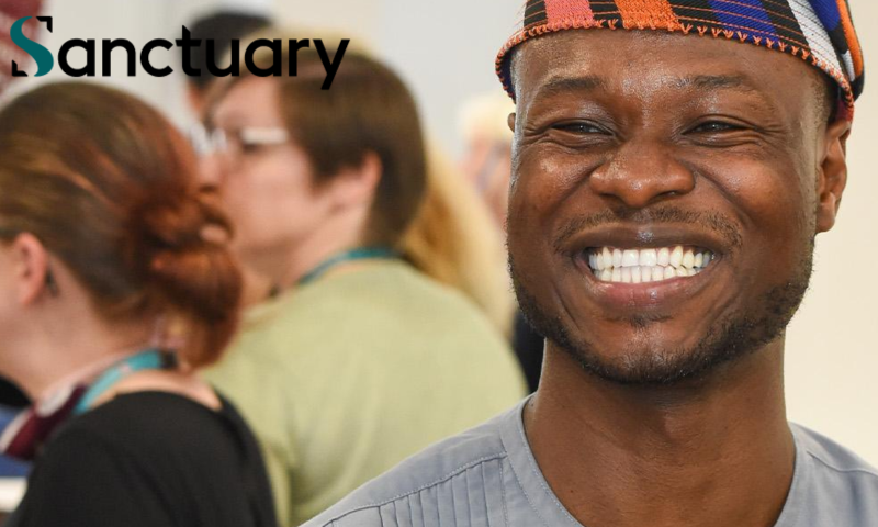 The image shows a man smiling broadly at the camera. He is wearing a colourful traditional West African cap, commonly known as a "fila" or Yoruba cap, featuring stripes of orange, blue, black and white. He is also wearing a grey traditional top that appears to be a dashiki or similar West African garment with vertical pleat detailing on the chest. The setting looks like an indoor social gathering or event, with several people visible but out of focus in the background.
