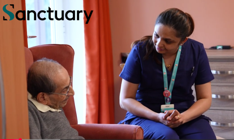 Key Elements of the Image The Healthcare Professional: On the right, a female carer is sitting and leaning forward slightly. She is wearing dark blue medical scrubs, a lanyard with an ID badge, and a pink badge. Her hair is tied back in a ponytail, and she has a kind, attentive expression as she looks down towards the resident. Her hands are gently clasped together in her lap.  The Resident: On the left, an elderly man is seated comfortably in a large, terracotta-coloured armchair. He is wearing glasses, a white-collared shirt underneath a grey jumper, and is looking up towards the carer with a calm expression.  The Setting: The environment looks cosy and domestic. In the background, there is a warm orange or terracotta curtain, soft pinkish-toned walls, and a wooden chest of drawers to the right.  Branding: In the top-left corner, the word "Sanctuary" is prominently displayed, featuring a distinct stylised teal "S" logo.