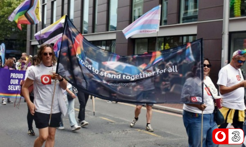 This is a photo of a Pride march taking place on a city street. A group of people are walking towards the camera carrying a large dark banner that reads "proud to stand together for all", with a smaller "#ProudTogether" hashtag beneath it. The banner has a faint photographic background and appears to be a VodafoneThree branded one. In the foreground on the left, a man in a white VodafoneThree t-shirt and shorts is holding one of the banner poles. He has light brown shoulder-length hair and is wearing sunglasses. Several other people in matching white t-shirts are carrying the rest of the banner and walking alongside it, including a person on the right using a long white cane. The background is full of Pride imagery: a Progress Pride flag and a Transgender Pride flag (light blue, pink and white) are flying on the left, and more rainbow and trans flags are visible on the right. To the left edge there's another purple banner, partly cut off, with white text that includes the words "PROUD" and "OUR" and "SELVES", suggesting something like "proud to be our [whole] selves". The setting is a paved road lined with modern buildings, with what looks like a balloon arch in the top right corner. The lighting is bright and overcast, and the overall mood is celebratory.