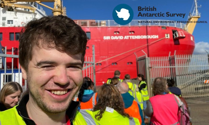 The Foreground Main Subject: On the left side of the frame, there is a close-up of a young man with brown, wavy hair and light stubble. He is smiling warmly at the camera. He is wearing a bright yellow high-visibility (hi-vis) safety vest over a black zip-up top.  Other Visitors: To his left (bottom-left corner), the top of a blonde woman’s head is visible. Behind him, a small crowd of people is walking away from the camera towards the ship. Several of them are wearing hi-vis vests in yellow and orange, while one person on the right wears a bright pink vest.  The Background The Research Vessel: Dominating the background is a massive, bright red ship. Printed on the side of the red hull in bold, white capital letters is the name "SIR DAVID ATTENBOROUGH".  The Environment: The ship is docked behind a grey metal security fence with vertical pales. Above the ship's hull, a yellow crane-like structure is visible against a clear, vibrant blue sky with a few faint clouds.  Branding In the top-right corner, there is a prominent overlay logo for the British Antarctic Survey (Natural Environment Research Council). The logo features a dark blue circular badge containing a white silhouette of the Antarctic continent, with the organisation's name printed alongside it in black text.