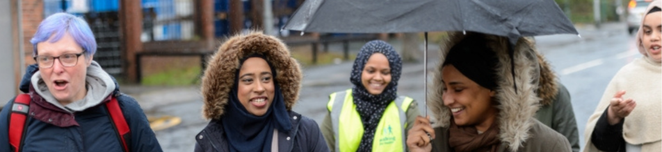 The image shows a diverse group of women walking together outdoors along a wet pavement on what appears to be a rainy or overcast day. Most of the women are smiling and engaged in friendly conversation as they walk side by side.  Detailed Breakdown Woman on the Far Left: Features vibrant, lilac-coloured hair, glasses, and a grey hooded top under a dark jacket with red backpack straps. She appears to be mid-speech.  Woman Second from Left: Wears a dark blue hijab paired with a dark winter coat that features a fluffy, faux-fur trimmed hood. She is smiling cheerfully toward the right.  Woman in the Centre (Background): Wears a dark patterned hijab and a bright, high-visibility yellow vest over her outfit. She is looking forward with a warm smile.  Woman Second from Right: Holds a large black umbrella overhead to shelter from the rain. She wears a brown scarf and a winter coat with a faux-fur hood, looking down with a happy expression.  Woman on the Far Right: Wears a cream-coloured headscarf and top, gesturing animatedly with her hands as she speaks to the group.  Setting & Atmosphere Weather & Environment: The ground is dark and wet, reflecting the damp weather. A dark umbrella is raised over the group to protect them from the rain.  Background: In the background, there is a brick wall/fenced area to the left, trees in the middle, and a wet road with traffic light reflections visible to the far right.  Tone: Overall, the mood is lively, warm, and communal despite the drab weather.