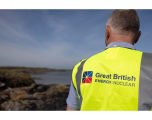 The image is an outdoor photograph taken from an over-the-shoulder perspective, looking past a person out towards a coastal landscape.  Foreground & Subject Person: Seen from behind, facing away from the camera. The individual has short, grey-and-salt-and-pepper hair and is wearing a light blue short-sleeved collared shirt beneath high-visibility gear.  Branded Vest: The person wears a bright high-visibility yellow vest featuring grey reflective vertical stripes on the sides. Printed across the centre back is the "Great British ENERGY | NUCLEAR" logo in dark blue text, alongside the geometric Union Jack-inspired emblem in blue, red, and yellow.  Background & Environment Coastal Landscape: The background shows a rugged, rocky shoreline leading into a body of water (sea or large estuary). The ground is dark, rocky terrain with patches of low-lying coastal vegetation.  Sky & Lighting: The sky is mostly clear blue with light, thin wisps of cloud. The natural daylight casts bright lighting across the back of the individual and the landscape beyond.