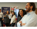 This image shows a candid, side-profile view of several individuals from the earlier Great British Nuclear group photo, standing together in an office setting and looking towards something off-camera with relaxed, smiling expressions.  Key Subjects (Foreground to Background) Man in Foreground (Right): Shown partially out of focus in the immediate foreground, standing with arms crossed, wearing a light blue button-down dress shirt and sporting a dark beard and moustache.  Woman in Sharp Focus (Centre): A woman with voluminous, dark curly hair smiling warmly. She is wearing a dark top under a textured grey jacket or blazer.  Woman in Grey Top (Mid-ground): Standing next in line with long brown hair, wearing a light grey long-sleeved jumper and dark trousers, also looking on and smiling.  Blonde Woman (Background): Slightly blurred, wearing a white long-sleeved top with blonde hair tied back or styled up.  Woman in Patterned Dress (Background): Standing further back, wearing a black-and-white patterned wrap dress with a blue lanyard around her neck, smiling towards the room.  Man on Far Left: Partially visible at the edge of the frame, wearing a light grey shirt with sleeves rolled up and dark trousers.  Setting & Background Branding: Behind the group stand the same dark blue pull-up banners bearing the "GREAT BRITISH NUCLEAR" header, alongside poster displays featuring various imagery and text.  Environment: A clean, brightly lit corporate room with neutral grey walls.
