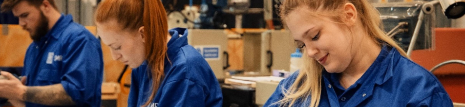 The image depicts three young apprentices working in an engineering workshop or technical training facility. •	Foreground (Right): A young woman with blonde hair tied back in a ponytail wearing bright blue work overalls (or a workshop jacket/shirt). She is looking down towards her work with a slight smile. •	Centre: A young woman with red hair tied back in a ponytail, facing left in profile. She is also clad in matching blue overalls and is intently focused on a task in front of her. •	Background (Left): A young man with dark hair and a beard, also wearing the same blue branded work shirt, looking down at his hands or equipment. The background shows industrial workshop equipment, machinery, and wooden partitions, bathed in warm lighting that gives the scene a focused, collaborative atmosphere.