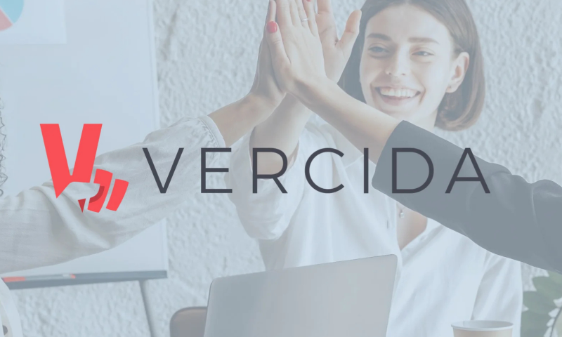 This image features the VERCIDA logo overlaying a high-five scene set in an office environment.  Branding: The logo is centered in the foreground, displaying the bright red stylized "V" symbol alongside the word VERCIDA in dark grey, uppercase lettering.  Background Scene:  People: A woman in a white blouse is smiling broadly in the background while high-fiving colleagues whose arms enter the frame from the left and right.  Environment: A bright, modern office space featuring a whiteboard with a pie chart visible in the top-left corner, an open laptop on a desk in the front, and a paper coffee cup.  As with the previous banner, the background photo is softly faded with a white overlay to keep the sharp logo branding front and centre.