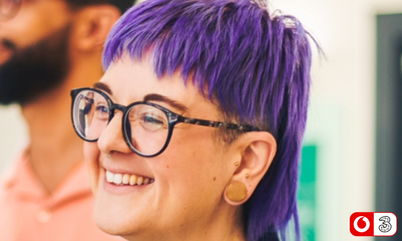 The image shows a close-up profile view of a smiling person with vibrant purple hair styled in a modern, textured mullet with short straight bangs. They are wearing round tortoise-shell, full-rimmed glasses, a subtle nose ring, and a large round flesh plug in their ear lobe alongside small tragus piercings.  They are smiling warmly while looking slightly off-camera to the left. In the background, the blurred figure of another person with a dark beard and a light peach-coloured shirt is partially visible. In the bottom right corner, there is a small logo featuring the VodafoneThree logo.