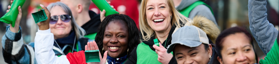 This image shows a cheerful group of supporters or volunteers outdoors, all smiling and actively celebrating.  Central Figures: At the centre of the group, a blonde woman in a black hoodie and bright green tabard or vest smiles widely while holding a green whistle to her mouth. Beside her on the left, a smiling Black woman holds up two small green noise-makers or clappers.  Surrounding Supporters: To the left, a woman in a patterned coat and sunglasses holds a green inflatable thunderstick high in the air. On the right, two more women are smiling toward the camera, one wearing a grey baseball cap and the other raising a arm wrapped in a striped long-sleeved top.  Branding & Colours: The group appears to be associated with a charity or event, wearing green tabards over their clothing and using bright green promotional noisemakers and inflatable clappers bearing white text.  Overall Atmosphere: The mood is lively, supportive, and energetic, characteristic of a cheering station at a marathon, fundraising event, or sports rally.