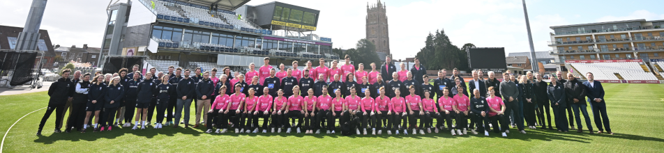 This panoramic wide-angle image shows a large group photo taken on the outfield of a cricket ground, featuring squad members, coaching staff, and officials posing together on bright green turf.  Foreground & Group Arrangement: A very wide ensemble of people is lined up across the pitch. The central block of players is wearing matching vibrant bright pink athletic shirts or jerseys, arranged in two main tiers—seated in the front row and standing directly behind them. Flanking this central group on both the left and right sides are coaches, backroom staff, and administrative personnel wearing dark tracksuits, jackets, and formal suits.  Background Architecture: Behind the team stands the main grandstand of the stadium, featuring white tiered seating, corporate hospitality boxes, and an electronic scoreboard above the main building structure.  Surroundings & Skyline: To the right in the background, a distinctive historic church tower rises into the clear sky, contrasting with the modern stadium stands and floodlight pylons.  Lighting & Atmosphere: The scene is brightly lit under direct sunlight with clear, light blue skies overhead.