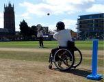 This image captures a wheelchair cricket match taking place at the Cooper Associates County Ground, the home of Somerset County Cricket Club (SCCC) in Taunton.  Foreground: A batsman seated in a specialized sports wheelchair faces towards the bowler. He wears a dark helmet, a white cricket shirt, and dark trousers, and holds a cricket bat mid-shot. To his right, in the bottom right corner, stands a set of bright blue plastic wickets.  Midground: A red cricket ball is suspended in the air between the batsman and an oncoming player in a white top and dark trousers, who appears to have just delivered the ball or is fielding on the pitch. An electronic scoreboard displaying match details stands prominently in the background.  Background: The iconic church tower of St James Church, Taunton, rises prominently on the left against a bright blue sky filled with light white clouds. To the right, modern pavilion buildings and spectator seating line the perimeter of the lush green outfield.