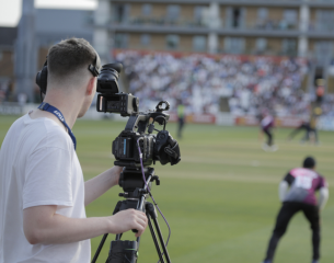This image shows a behind-the-scenes view of a camera operator filming a live cricket match.  Foreground: A videographer, seen from behind wearing a white t-shirt, headphones, and a lanyard labeled "STAFF", operates a professional broadcast camera mounted on a tall tripod.  Midground: To the right, a cricket player wearing a dark kit with pink details is positioned in the field on the boundary edge, facing away from the camera.  Background: The cricket pitch and outfield stretch out towards a crowded grandstand packed with spectators, set against the backdrop of the stadium architecture, all bathed in natural daylight.