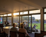 This image shows guests viewing a match from an indoor hospitality suite or lounge overlooking the ground.  Foreground: Several spectators sit indoors with their backs to the camera, looking out towards the pitch. In the room, round dining tables laid with white tablecloths and numbered cards are visible.  Midground: Large floor-to-ceiling glass doors and windows run the length of the room, separating the indoor dining area from an outdoor balcony seating section, where spectators are watching the play. Bright sunlight streams through the glass, creating a warm glow.  Background: Beyond the glass, the green cricket pitch, grandstands packed with crowd members, and the opposite stadium pavilion buildings are clearly visible under a late afternoon sun.