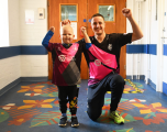 This image shows a young child and an adult man posing together indoors, both wearing Somerset County Cricket Club training tops featuring the Trade Nation sponsor logo.  Subjects: On the left, a young child stands triumphantly raising both fists in the air, wearing a pink, black, and blue kit over dark trousers. Beside the child, a smiling man kneels on one knee, raising his left arm in a matching victory pose. He wears a black Somerset CCC shirt, dark tracksuit bottoms, and bright yellow-green trainers.  Setting: They are positioned in a corridor with light-coloured brick walls, a blue dado stripe, double wooden doors behind them, and a distinctive patterned vinyl floor decorated with colorful leaf and floral designs.