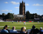 This image shows a County Championship cricket match underway at the Cooper Associates County Ground in Taunton, framed by its iconic landmark background.  Foreground: The blurred backs of several spectators sit in the boundary seating, watching the match unfold on the field.  Midground: On the bright green cricket pitch, players clad in traditional white cricket whites are active during play. An fielder is positioned in the center, whilst players, umpires, and wickets are dotted across the square.  Background: The tall, red-sandstone church tower and tiled roof of St James Church stand prominently behind the ground perimeter under a bright blue, cloud-dotted sky. To the right of the church, a large electronic scoreboard displays live match details for a fixture between Somerset and Surrey.