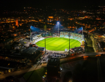 This aerial night photograph shows an illuminated Cooper Associates County Ground, home of Somerset County Cricket Club in Taunton.  Center: The brightly lit oval green cricket pitch acts as the focal point, illuminated by towering floodlights whose beams cut through the dark sky.  Immediate Surroundings: Surrounding the pitch are the stadium's pavilions, grandstands, and adjacent car parks filled with vehicles.  Background: Beyond the venue, the glowing streetlights and buildings of the town of Taunton stretch across the dark landscape, extending toward the horizon.