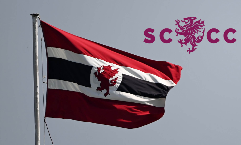 This image shows the flag of Somerset County Cricket Club flying against a pale, overcast sky, alongside an overlaid club logo in the top-right corner.  The Flag: A large woven flag attached to a grey metal flagpole on the left side, fluttering in the wind. It features wide top and bottom horizontal bands of deep crimson/maroon, sandwiching a white middle field. Passing through the white centre is a thick black horizontal stripe, which is interrupted in the middle by a red wyvern emblem.  Top-Right Overlay: The official Somerset County Cricket Club logo superimposed onto the sky. It comprises bold maroon letters "SC" and "CC" separated by a detailed graphic of a maroon wyvern standing upright.