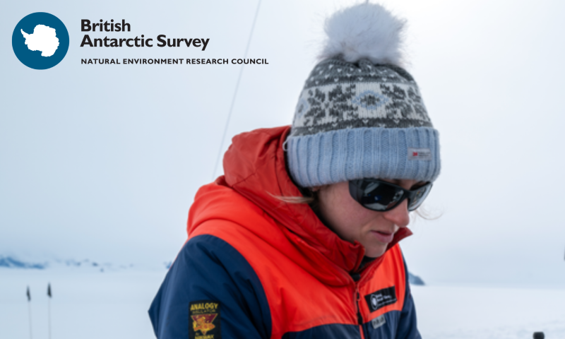 photo of Emma Fisher, now set up as a British Antarctic Survey banner. She is shown in profile, looking down at her work, wearing a navy and orange insulated jacket, a grey and blue knitted bobble hat and dark sunglasses. Behind her is a flat expanse of snow and ice under a pale sky, with marker flags and faint hills on the left. The British Antarctic Survey logo and "Natural Environment Research Council" strapline sit in the top-left corner.