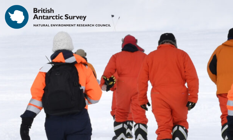 A group of people in bright orange cold-weather gear walk away from the camera across flat snow, seen from behind. On the left, one wears an orange hi-vis jacket, navy trousers, a white bobble hat and a black backpack. The others are in full orange insulated boiler suits with heavy boots, hats and gloves, spread out in a loose line as they head towards a marker flag on the horizon. The sky is pale and overcast. The British Antarctic Survey logo and "Natural Environment Research Council" strapline sit in the top-left corner as part of the banner layout.