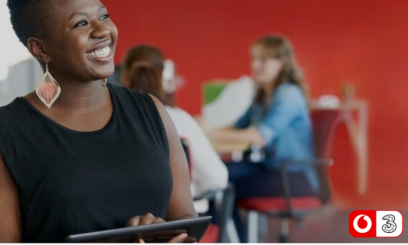The image shows a smiling Black woman with short hair in a modern office environment. She is positioned in the foreground, turned slightly to the right, looking off-camera with a warm expression. She is wearing a sleeveless black top, large woven diamond-shaped drop earrings in a coral/peach tone, and holding a tablet computer in her hands.  In the background, which is softly blurred, there is a vibrant red wall and two other colleagues sitting at a table engaged in conversation. One person has long brown hair viewed from behind, while the other is wearing a blue denim shirt.  In the bottom right-hand corner, there is a logo showing the Vodafone speech mark inside a red circle alongside the Three UK logo.