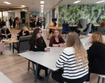 Foreground GroupSeating: Four women are gathered around a large, grey rectangular table supported by dark blue A-frame legs.   Left Side: A woman with dark wavy hair and glasses wears a black top, sitting with arms crossed. Next to her, a woman in a burgundy long-sleeved top and a blue smartwatch holds a paper takeaway coffee cup.   Right Side: In the foreground with her back to the camera, a woman with long blonde hair wears a white jumper with black horizontal stripes. Beside her sits a woman in a dark jacket or cardigan with shoulder-length blonde hair.   Background & EnvironmentWall Feature: The back-right wall features the same green tropical leaf-patterned wallpaper seen in the previous image, complete with hanging potted plants.   Booths & Seating: The high-backed privacy booths from the first picture are visible against the featured wall on the right. Blue accent armchairs and grey soft seating are dotted throughout the space.   Open Layout: To the left, there is a lounge area with low tables, armchairs, and timber structural frames overhead. Groups of colleagues are chatting and networking in the distance, giving the office a busy, community-focused feel.   Finishes: The space combines light wood-effect laminate flooring in the central path with carpeted seating zones and recessed grid lighting along the ceiling.