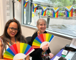 Main SubjectsWoman on the Left: Has long dark hair, black-framed glasses, and red lipstick, wearing a patterned sleeveless top and a rainbow-coloured lanyard. She is holding up a unfolded paper hand fan displaying a full rainbow spectrum.   Woman on the Right: Has blonde hair styled with a fringe, glasses, and is wearing a black top with a colourful lanyard. She holds a matching rainbow-patterned hand fan.   Decor & BackgroundBunting: Progress Pride flag bunting (featuring the classic rainbow with the intersex-inclusive chevron design) is strung across the top of the window frame behind them.   Setting: They are sitting in front of a large window looking out onto a paved outdoor area and car park. UK standard triple light switches or sockets are visible on the white wall to the far left.   Desk setup: On the right foreground, a blue-covered table holds a laptop, phone, pencil case, and printed paperwork.