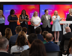 Presenters on StageStanding in front of two large presentation screens displaying a red and blue gradient background with the text "Question time.", there are seven panel members (left to right):   Woman on far left: Wearing a light blue striped button-down shirt and black trousers.   Second woman: Wearing a sleeveless, navy blue midi dress with a square neckline.   Third woman: Wearing a navy sleeveless top with a patterned neck scarf.   Fourth woman: Wearing a white short-sleeved top with a red name badge, holding notes.   Man: Wearing a light grey patterned/plaid blazer over an open-collar white shirt and dark trousers.   Fifth woman: Standing in a light pastel blue knee-length dress, holding a microphone and addressing the room.   Woman on far right: Wearing a dark tailored blazer/suit, standing behind a white podium.   Audience & SettingAudience: The foreground shows the backs of numerous seated audience members listening to the presentation.   Setup: A professional conference or auditorium setting with a metal truss stand supporting the podium and dual-screen digital displays mounted on the wall behind the speakers.