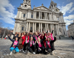 Group & AttireParticipants: A diverse group of roughly 20 people are posing enthusiastically for a group photo, waving, cheering, and raising their hands in the air.   Clothing: Many participants are wearing bright pink t-shirts featuring the Refuge charity logo, styled over coats, jackets, or jumpers. Others wear red coats, gilets, or smart casual outerwear.   Background & SettingLocation: The iconic Grand Western Facade of St Paul’s Cathedral dominates the background, showcasing its classic Baroque architecture, tall stone columns, twin bell towers, and detailed pediment.   Environment: The group is standing on the paved stone plaza in front of the cathedral steps under a partly cloudy blue sky. Other pedestrians and bystanders can be seen in the distance around the square.