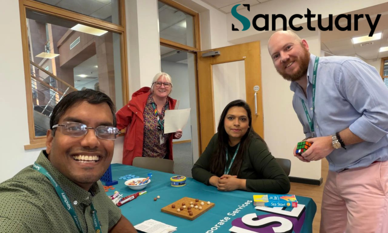 This photograph shows four colleagues in an office setting, gathered around a table setup for an interactive event or workshop. The word "Sanctuary" is prominently displayed in large text in the upper right corner.   People and Positions:Foreground Left: A smiling man taking a selfie, wearing glasses, a patterned green short-sleeved shirt, and a teal "Sanctuary" lanyard.   Background Center-Left: A woman with white hair and glasses wearing a red jacket over a floral top, smiling while holding a sheet of paper.   Center: A woman with long dark hair wearing a dark green long-sleeved top and a teal lanyard, seated at the table with her hands clasped.   Foreground Right: A smiling bald man with a beard, wearing a light blue button-down shirt with rolled-up sleeves, light pink trousers, and a teal lanyard. He is standing while holding a Rubik's Cube.   Table Setup:The table is covered with a teal tablecloth featuring white and purple lettering along the front edge that reads "porate Services" (part of "Corporate Services").   Displayed on the table are various games and refreshments, including a small wooden solitaire/marbles board game, a Connect Four set, a bowl of sweets, wrapped snacks, and various card games or activity boxes (one labeled "Sea Shanties").   Setting and Background:The scene takes place in a bright, modern office with light-coloured walls, wooden door frames, and large interior windows looking out onto a hallway and staircase.