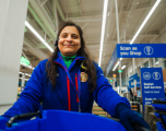 Subject: A female staff member wearing a blue Tesco fleece jacket with red trim over a green high-neck top. She is wearing a Tesco lanyard around her neck, a name badge, a headset, and dark gloves. She is positioned behind a bright blue plastic shopping crate or trolley bin.   Foreground: The top rim and handles of a blue crate dominate the immediate lower foreground. Stacked blue shopping baskets are also visible to the right.   Background: The store interior features high industrial ceilings with bright overhead strip lighting. Blue Tesco signage is visible in the background, including a sign reading "Scan as you Shop" with an icon of a handheld scanner, and another sign for "Basket Self Service Cash & Card". A green store department sign is softly blurred in the distance to the left.   Composition & Lighting: The image is shot from a low angle, giving a clear view of the ceiling structure and signage overhead. The lighting is vibrant and typical of modern supermarket interiors.