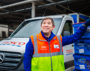 Subject: A male colleague dressed in a long-sleeved blue Tesco fleece layered under a high-visibility hi-vis vest. The vest is orange on top and neon yellow-green on the bottom with silver reflective strips, featuring the branded Tesco logo and "Every little helps" slogan on the chest, along with an ID badge. He has one hand resting on a stack of delivery crates and is looking up and slightly away from the camera with a cheerful expression.   Vehicle & Crates: Behind him is a white Mercedes-Benz Tesco delivery van featuring the red and blue Tesco logo on the bonnet and side. To the right is a tall stack of blue plastic delivery crates filled with grocery items, complete with white barcode labels attached.   Background & Lighting: The setting appears to be a loading bay or depot yard under an overhang or covered structure. The image uses a natural light focus with a shallow depth of field, keeping the driver in crisp focus while softly blurring the background vehicle.