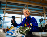 This photograph depicts a smiling female Tesco checkout colleague operating a till inside a store.   Subject: An older female staff member with short grey hair and glasses, wearing a blue Tesco fleece jacket with red details. She has a circular Tesco badge pinned to her chest and is holding a black handheld barcode scanner in her right hand.   Foreground: A conveyor belt area featuring grocery items, including a bag of salad greens, bananas, and a melon. Part of a till screen is visible on the far left.   Background: The checkout till is set against large glass window panels with timber frames, letting in natural daylight. An ATM sign and additional till monitors are faintly visible in the background.   Composition & Lighting: Shot from a low, slightly angled perspective near the checkout counter, capturing bright overhead lighting combined with soft daylight streaming through the windows.