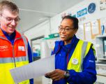 Subjects:On the right, a female colleague named Natasha is wearing a blue Tesco fleece jacket layered under a neon yellow high-visibility vest. She wears tortoiseshell glasses, a digital wrist watch, and a white Tesco name badge. She is smiling as she holds up a sheet of printed paper.   On the left, a male colleague wearing glasses and a high-visibility vest (orange top, yellow bottom with silver reflective striping) over a blue Tesco shirt looks down attentively at the document with a smile.   Foreground: The printed sheet of paper held between the two colleagues forms the central focal point.   Background: An office setting with white walls, a round wall clock, noticeboards pinned with information sheets, and computer equipment.   Composition & Lighting: The image features a bright, candid composition shot at eye level, highlighted by clean overhead commercial lighting.