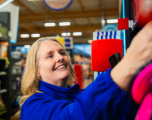 This photograph shows a smiling female Tesco colleague filling or arranging stock on a store shelf.   Subject: A blonde female staff member wearing a high-collared, long-sleeved blue Tesco jacket. She is smiling cheerfully as she reaches up towards a display on the right-hand side. She appears to be holding a handheld barcode terminal or mobile device in her hand as she works.   Foreground: The right side of the image features brightly coloured red and pink packaging on a retail display shelf, partially blurring into the foreground.   Background: The background shows a typical supermarket aisle with warm wooden ceiling beams and bright overhead strip lighting. Blurred shelf units, stock displays, and hanging in-store promotional signage are visible further down the aisle.   Composition & Lighting: Shot from a low, close-up side angle focusing closely on the staff member's expression, utilizing a shallow depth of field to keep her sharp while softly blurring the aisle behind her.