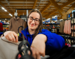 This photograph shows a smiling female Tesco colleague arranging clothing on a clothing rack in the store's F&F apparel department.   Subject: A young female staff member with long dark hair, wearing black glasses, a headset, and a blue Tesco fleece jacket with red trim. She is smiling as she handles and organises garments on clothes hangers.   Foreground: A close-up view of dark plastic coat hangers and light-coloured fabric items on the rack she is working on.   Background: The clothing section features various items of clothing displayed on hangers, including jumpers and tops on surrounding racks. The high ceilings feature wooden support beams and bright retail overhead lighting, with blue promotional signs visible in the distance.   Composition & Lighting: The shot is captured at a slightly low, medium close-up angle, using soft daylight and bright interior store lighting with a shallow depth of field to keep the focus firmly on the staff member.