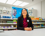 Subject: A female staff member with dark, shoulder-length hair, wearing a patterned red and purple top under a black blazer. She has her hands clasped on the counter and is wearing a circular white Tesco badge that reads "Rita, Pharmacy Manager".   Foreground: A clean, white pharmacy service counter fills the bottom of the frame.   Background: Shelving units stocked with various over-the-counter health and medicinal products displaying clear price labels (e.g. £5, £8.75, £12, £40). A large digital display screen behind her shows a toothpaste or oral health promotional advertisement featuring a smiling child.   Composition & Lighting: Shot from a low, medium angle over the counter, illuminated by bright, clean overhead light panels embedded in the white ceiling.
