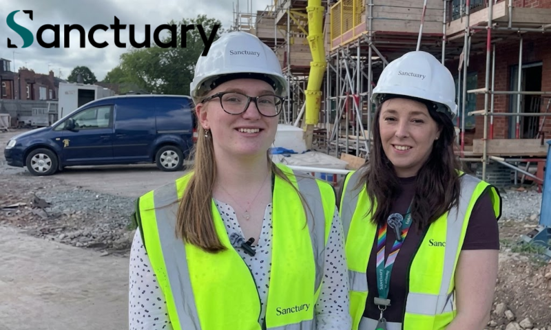 The image depicts two women smiling side by side outdoors on an active construction site, both wearing personal protective equipment (PPE).  The Subjects:  Person on the left: A woman with long blonde hair and dark-rimmed spectacles, smiling towards the camera. She wears a white safety hard hat branded with the "Sanctuary" logo, a white blouse patterned with small dark spots, and a fluorescent yellow high-visibility waistcoat with reflective silver strips and "Sanctuary" printed on the lower front. A small clip-on wireless microphone is attached to her neckline.  Person on the right: A woman with dark brown, shoulder-length hair, also smiling. She is wearing a matching white "Sanctuary" hard hat, a dark plum-coloured T-shirt beneath a fluorescent yellow high-visibility vest, and a rainbow lanyard around her neck holding an ID badge, to which a lapel microphone is clipped.  Background and Setting:  Behind them is an active residential building development. To the right stands a multi-storey brick building surrounded by metal scaffolding, complete with safety netting and a bright yellow vertical rubble chute.  To the left, a dark blue van is parked on the unpaved site ground, alongside site cabins and traditional terraced houses visible in the far distance under an overcast sky.  Branding:  A prominent "Sanctuary" corporate logo in teal and black is overlaid in the upper-left corner of the frame.