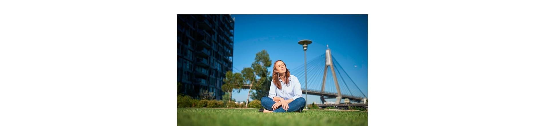 wellbeing at lendlease Woman sat in a field with green grass and blue sky with a building in the background