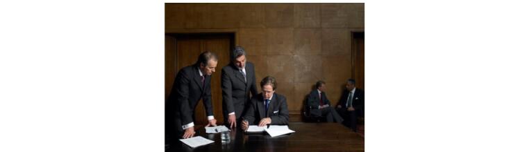 Group of men in suits stood and sat around a table with papers in front of them