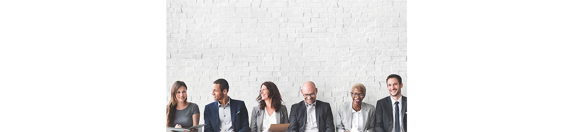 a diverse group of people sitting next to each other, with their backs against a white brick wall. They are speaking to and laughing with each other. 