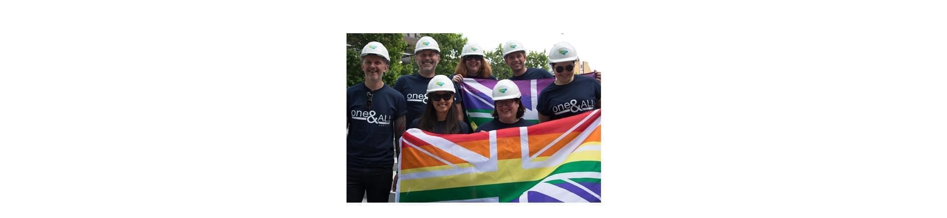 Lendlease is the Second Industry Firm to Meet the Equality Standard a group of seven Lendlease employees wearing Lendlease branded hard hats. They are holding British rainbow flags and wearing tops that read 'One and All'.